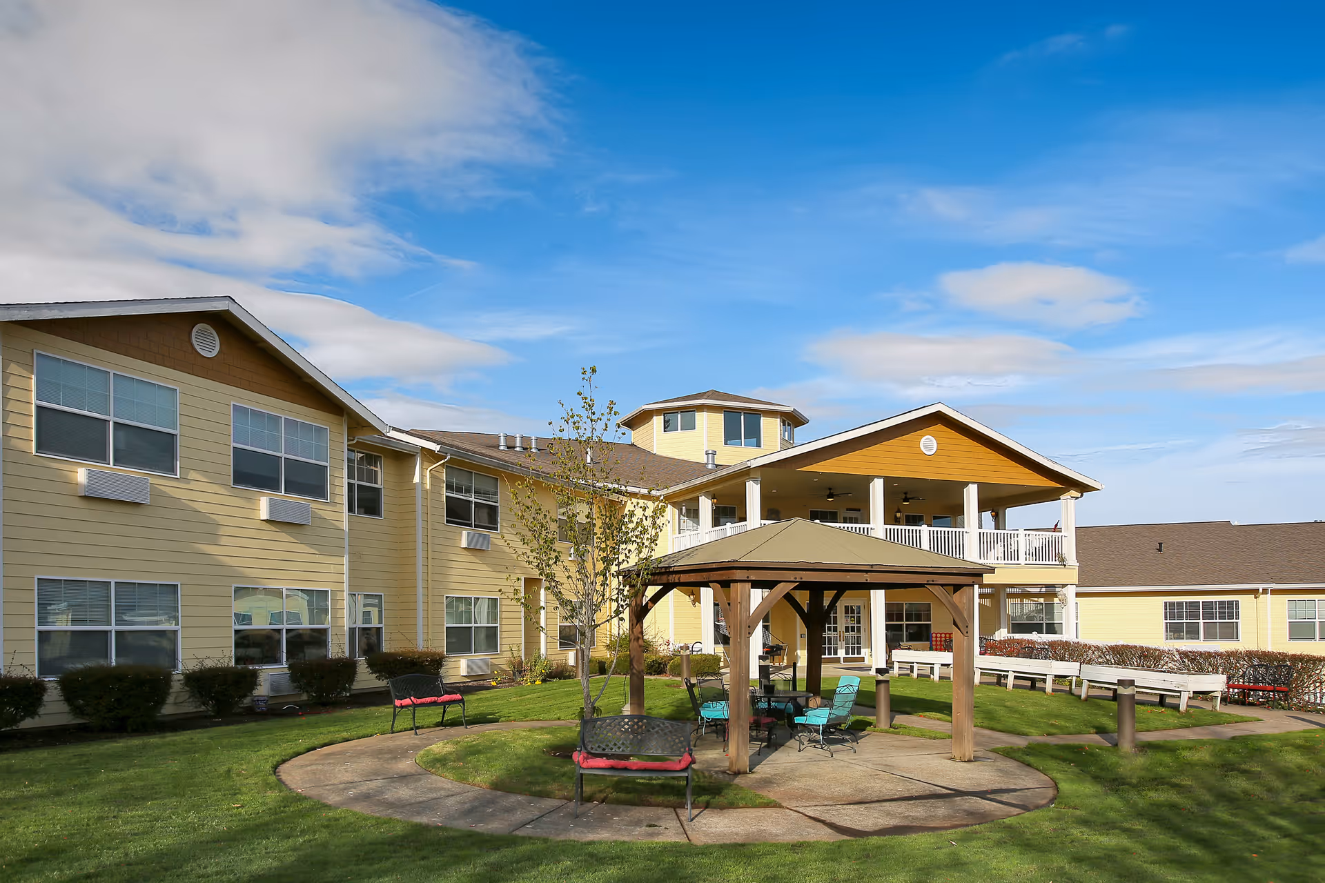 Outdoor view of a senior living facility with a yellow two-story building, a covered gazebo with seating, benches, and a well-maintained lawn under a partly cloudy blue sky.