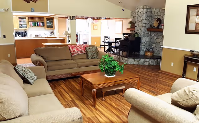 A cozy living room area with three beige sofas arranged around a wooden coffee table with a green potted plant on it. The room has wooden flooring, a stone fireplace in the corner, and an open view into a dining area and kitchen in the background.