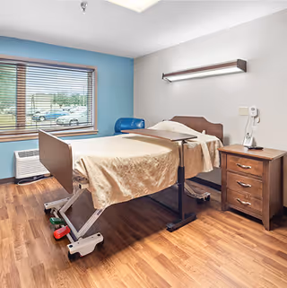 A senior living facility bedroom with a hospital-style adjustable bed covered with a beige blanket. The room has wooden flooring, a wooden nightstand with drawers, a wall-mounted light above the bed, and a window with blinds showing a parking lot outside.