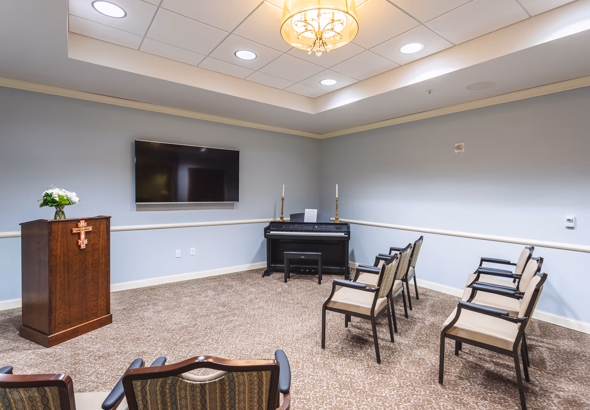 A small chapel or meeting room with a wooden podium adorned with a cross and flowers on the left, a digital piano with two candlesticks and sheet music against the far wall, a flat-screen TV mounted on the wall, and several chairs arranged in rows facing the podium and piano. The room has light blue walls, a patterned carpet, and a chandelier hanging from the ceiling.