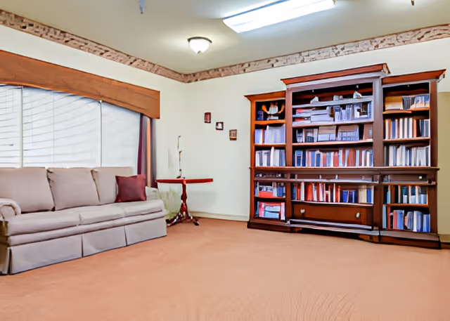 Living room with a beige sofa, wooden bookshelf full of books, a small side table, and a large window with blinds.