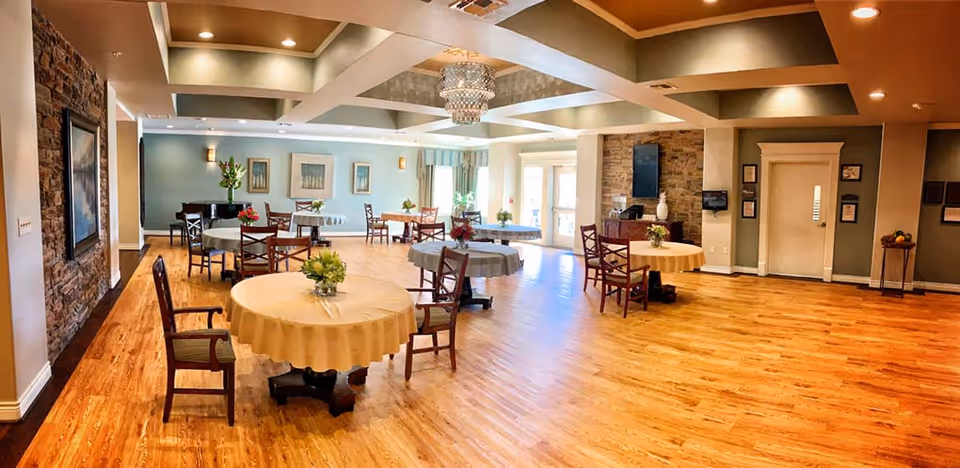 Spacious dining room with round tables covered in tablecloths, each adorned with a small floral centerpiece. Wooden chairs surround the tables. The room features wooden flooring, a coffered ceiling with recessed lighting, a stone accent wall with a mounted TV, and large windows letting in natural light. There is a piano in the corner and framed artwork on the walls.