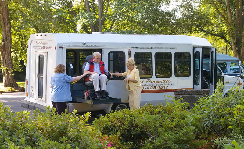An elderly man in a motorized wheelchair is being assisted by two women as he is lifted onto a white shuttle bus. The scene is outdoors with green trees and bushes surrounding the area.