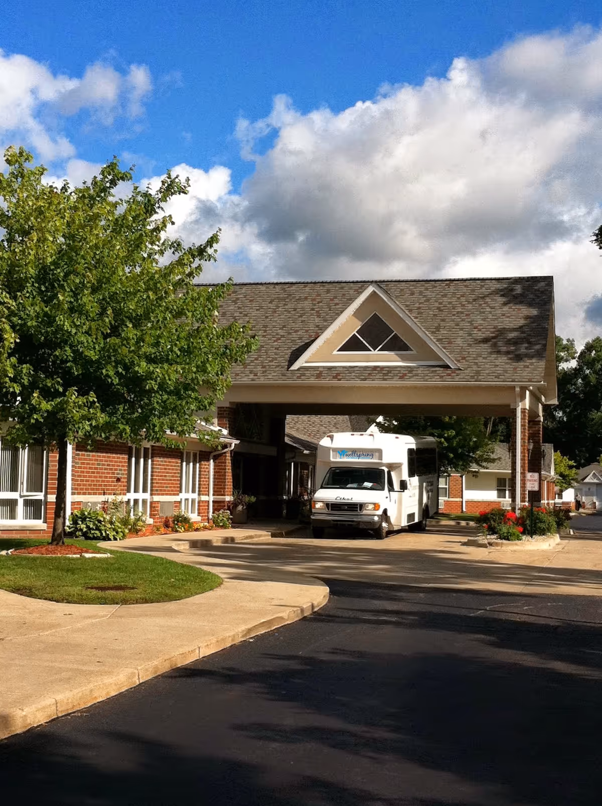 Exterior view of Wellspring Lutheran Services building with a covered entrance. A white shuttle bus is parked under the entrance canopy. There is a tree and landscaped greenery on the left side, and a clear blue sky with some clouds above.