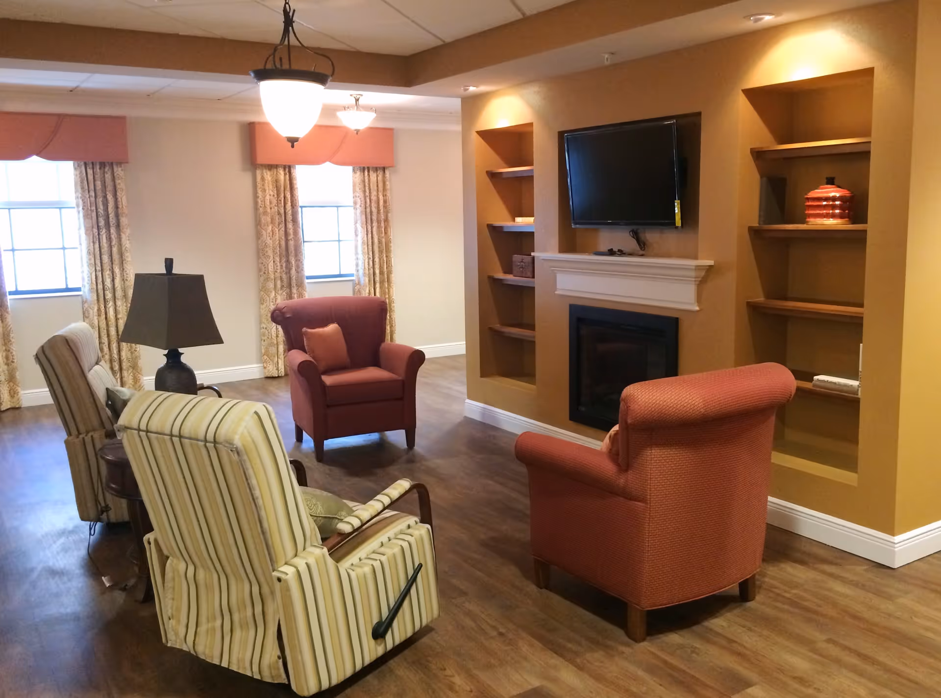 A cozy living room area with four armchairs arranged around a fireplace and a wall-mounted flat-screen TV. The room has wooden flooring, built-in shelves on either side of the fireplace, two windows with patterned curtains and valances, and a ceiling light fixture.