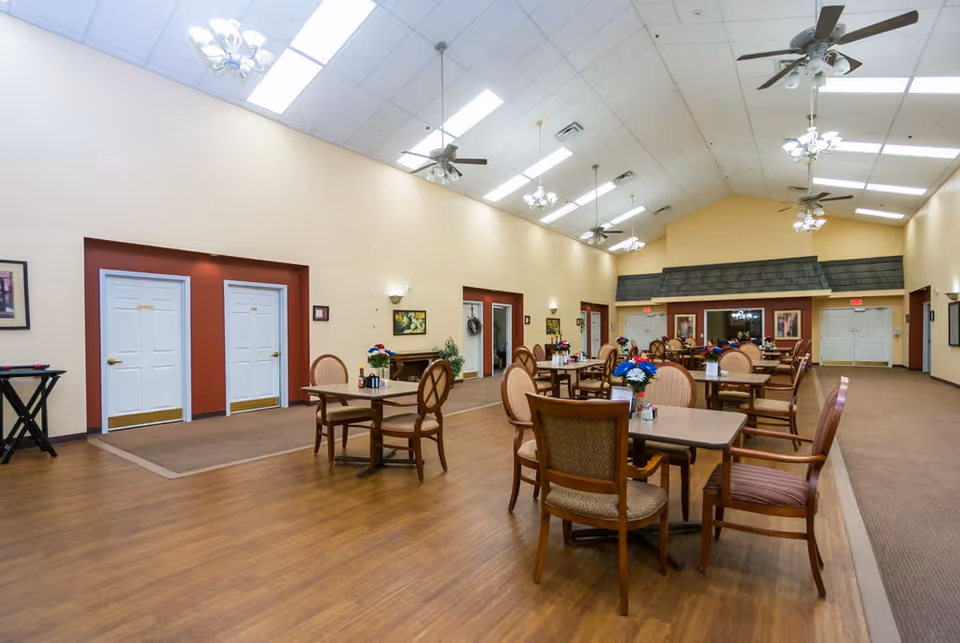 A spacious dining area with multiple tables and chairs arranged neatly. The room has a high ceiling with ceiling fans and hanging light fixtures. The walls are painted beige with some red accent walls around doorways. There are framed pictures and plants decorating the walls. The floor is a combination of wood and carpet. The room appears well-lit with natural and artificial lighting.
