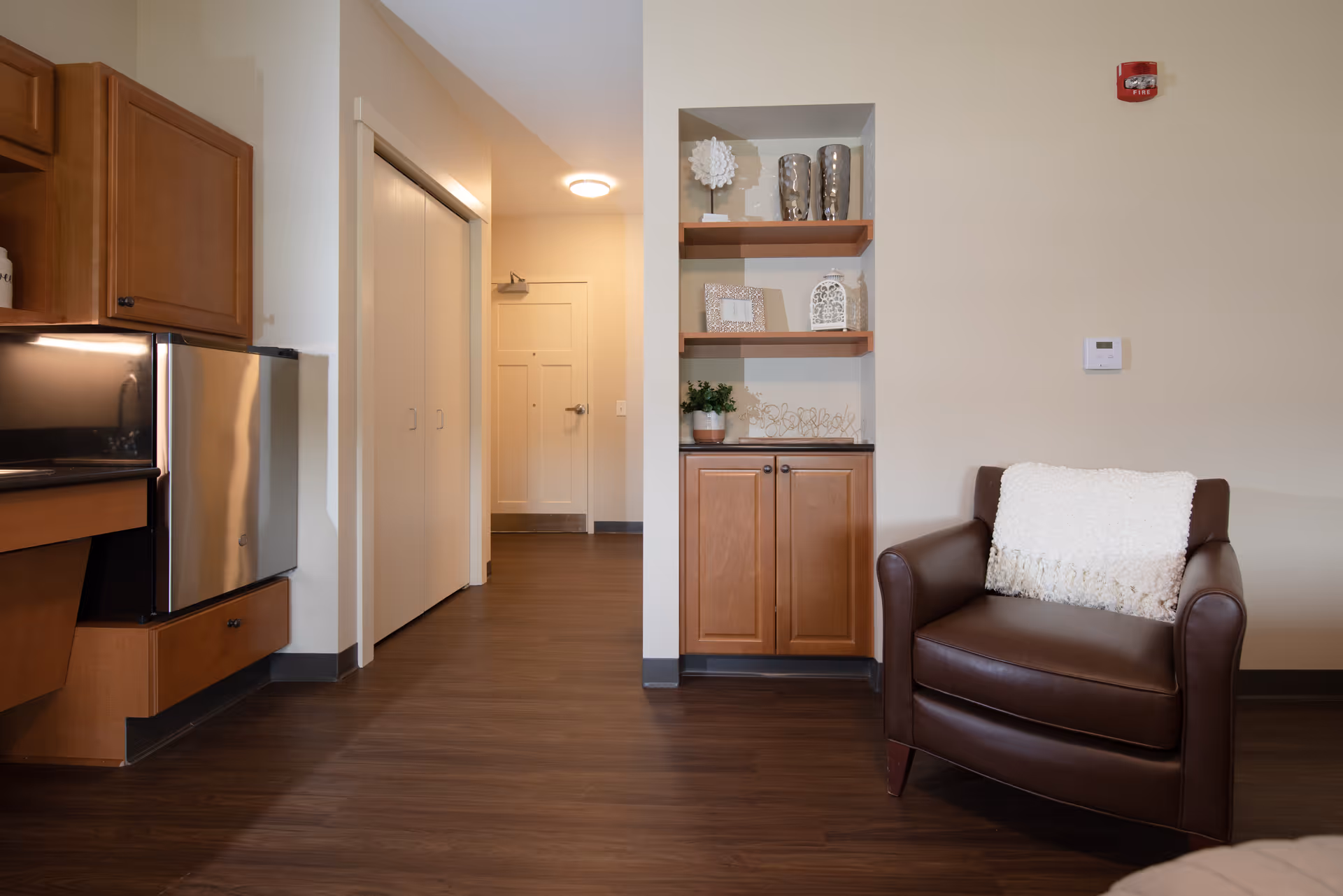Interior view of a senior living facility room showing a brown leather armchair with a white throw blanket, wooden shelves with decorative items, a small cabinet, and part of a kitchen area with wooden cabinets and a stainless steel mini fridge. The floor is dark wood, and there is a hallway leading to a closed door in the background.