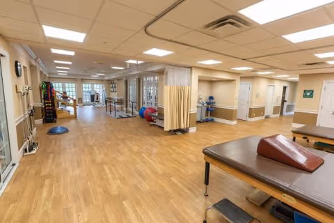 A spacious rehabilitation or physical therapy room with wooden flooring, therapy tables, exercise equipment including balance balls and parallel bars, and a privacy curtain. The room is well-lit with ceiling lights and has beige walls with white trim.