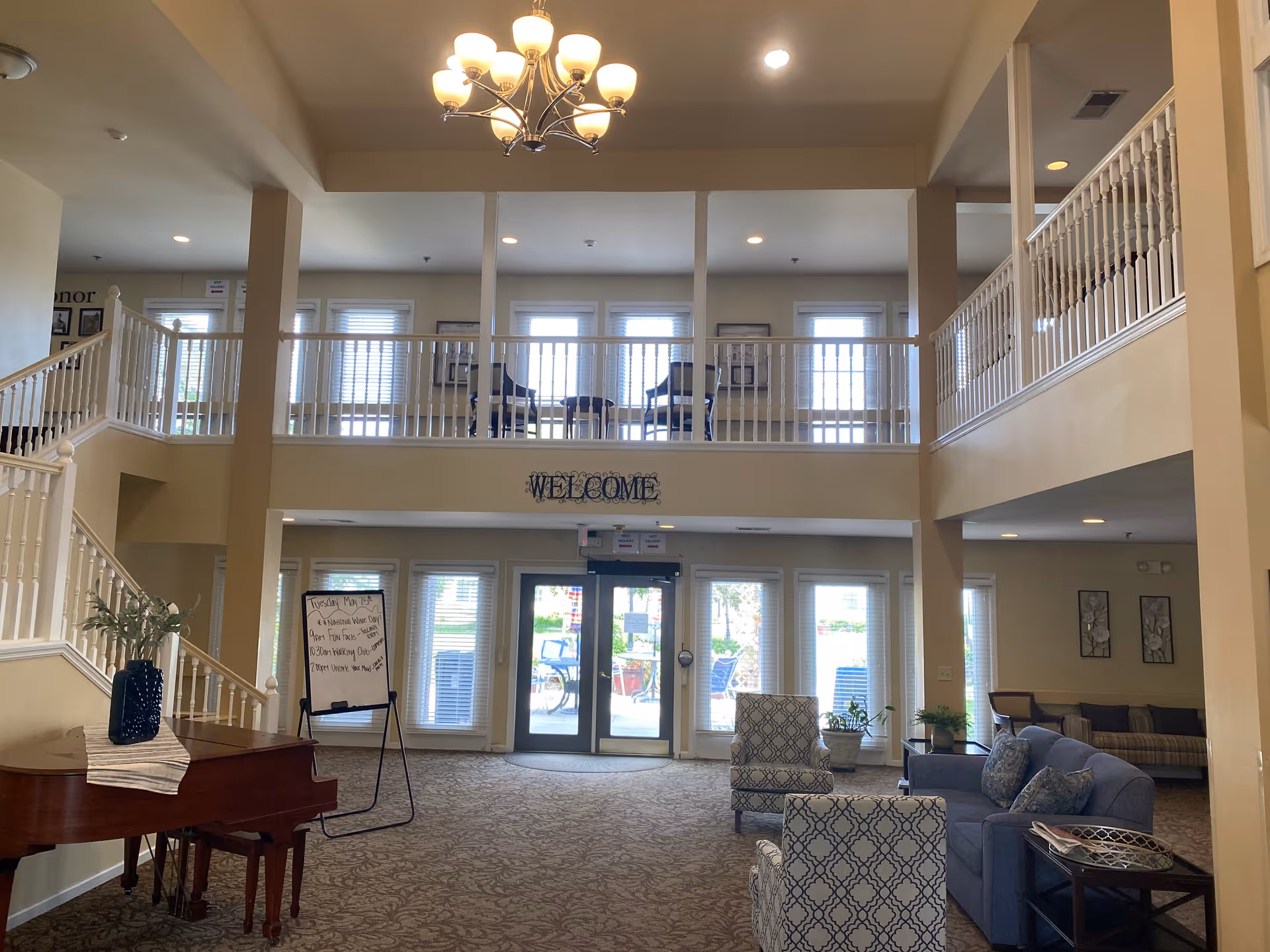 Spacious senior living facility lobby with high ceilings and a balcony overlooking the entrance. The area features a grand piano with a decorative vase, comfortable seating including patterned armchairs and a sofa, and a welcome sign above the main entrance doors. Large windows allow natural light to fill the space.