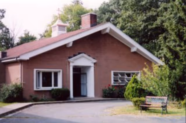 A single-story brick building with a peaked roof and a small cupola on top, surrounded by trees and bushes. There is a paved driveway leading to the entrance, which has a white door frame with a small pediment above it. A bench is placed on the grass near the driveway.