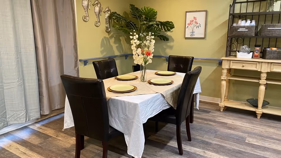 A dining area with a rectangular table covered with a white tablecloth and a beige runner, set with four plates. Four dark brown leather chairs surround the table. A vase with white and red flowers is placed in the center. The room has light green walls, wooden flooring, a sideboard with baskets and cups, and decorative seahorse wall hangings.