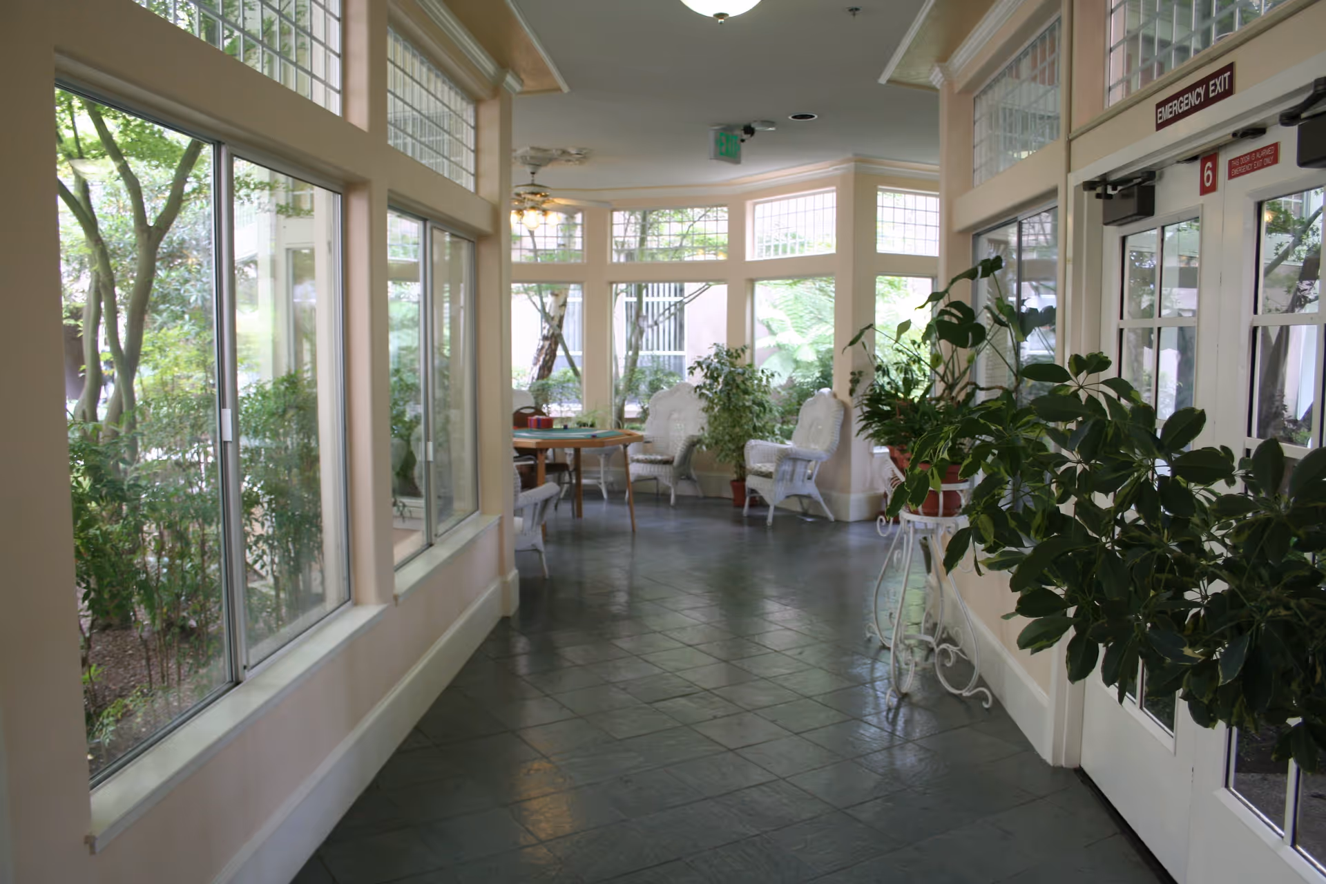 A bright indoor hallway with large windows on the left side showing greenery outside. The hallway has a tiled floor and is decorated with several potted plants. At the end of the hallway, there are white wicker chairs and a round table with chairs around it. On the right side, there is a glass door with an emergency exit sign above it.