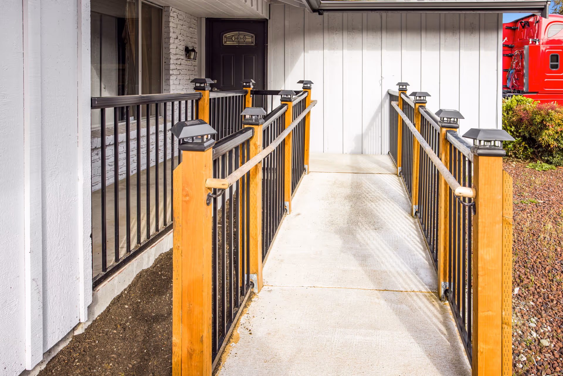 A concrete wheelchair accessible ramp with wooden posts and black metal railings leading to a dark-colored door on the exterior of a white building. There are small solar lights mounted on top of the wooden posts and some bushes and a red truck visible in the background.
