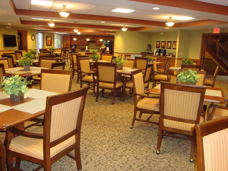 A spacious dining room with multiple wooden tables and cushioned chairs arranged neatly. Each table has a small green plant centerpiece. The room has warm lighting with ceiling fixtures and recessed lights. There is a carpeted floor with a patterned design, and the walls are painted in soft yellow and beige tones. In the background, there is a counter area with coffee machines and framed artwork on the wall, and a staircase is visible to the right.