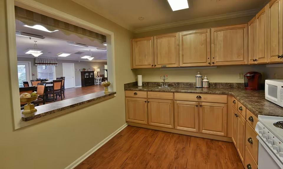Interior view of a kitchen with wooden cabinets, a countertop with a sink, a microwave, and a coffee maker. There is a pass-through window looking into a dining area with tables and chairs, hardwood flooring, and ceiling lights.
