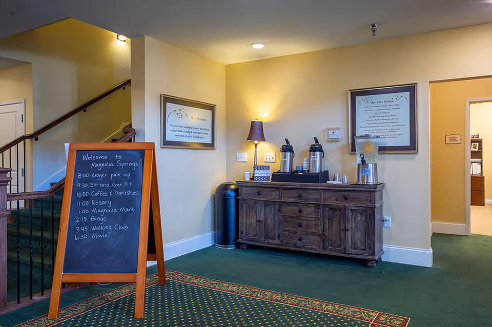 Interior view of a senior living facility lobby area with a wooden sideboard holding coffee dispensers, cups, and a water dispenser. A floor lamp is on the sideboard, and two framed notices hang on the wall behind it. A chalkboard sign lists daily activities including Kroger pick up, Sit and Get Fit, Coffee & Danishes, Rosary, Magnolia Mart, Bingo, Walking Club, and Movie. There is a staircase with a wooden handrail and green carpet leading upstairs, and an open doorway to another room.