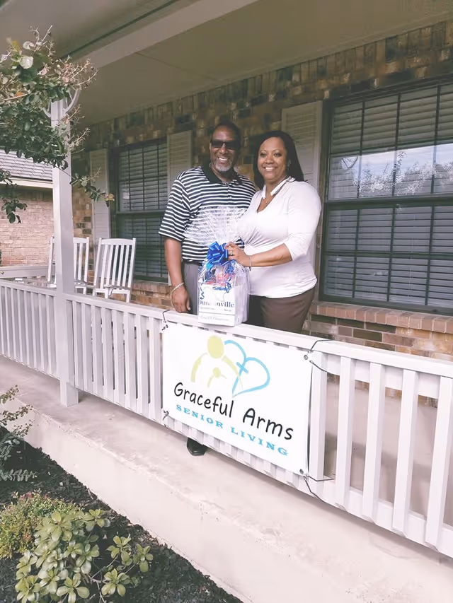 Two smiling adults stand on a front porch behind a railing with a 'Graceful Arms Senior Living' sign, holding a wrapped gift basket.