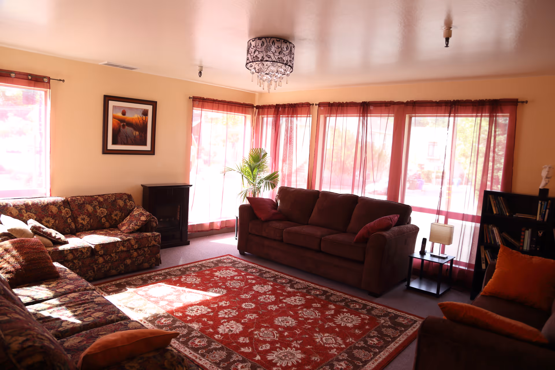 A cozy living room with three sofas arranged around a large red and beige patterned area rug. The room has large windows with sheer red curtains allowing natural light to fill the space. There is a small black electric fireplace, a side table with a lamp and a phone, a bookshelf with books and a decorative bust, and a framed landscape painting on the wall. A decorative ceiling light fixture hangs from the ceiling.