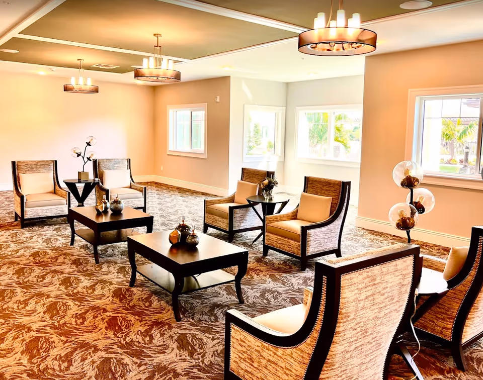 A bright and spacious living room area with multiple cushioned armchairs arranged around dark wooden coffee tables. The room has large windows letting in natural light, patterned carpet flooring, and modern ceiling light fixtures. Decorative items are placed on the tables and a unique floor lamp with three glass globes stands near the window.