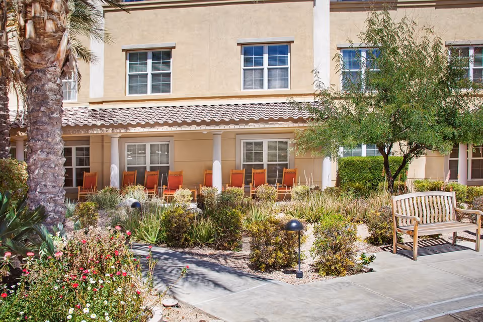 Outdoor patio area at a senior living facility with a row of orange chairs lined up under a covered porch, surrounded by landscaped bushes, flowers, a palm tree, and a wooden bench on a concrete pathway.
