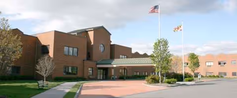 Brick nursing home exterior with entrance canopy, flagpoles, and surrounding trees.