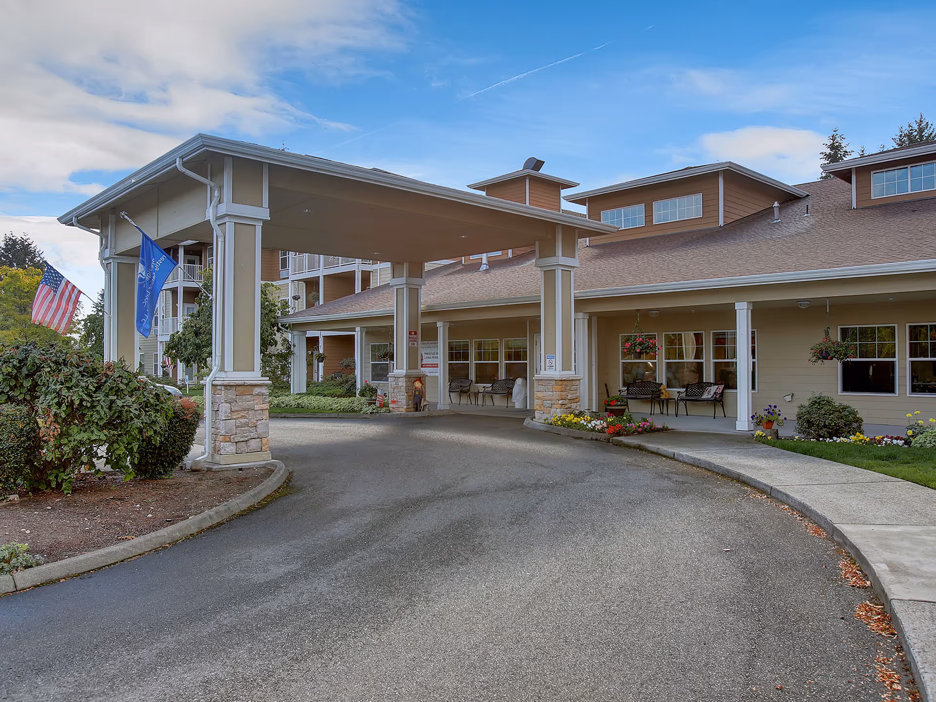 Covered entrance/porte-cochere of a senior living building with flags, seating, and a landscaped driveway.