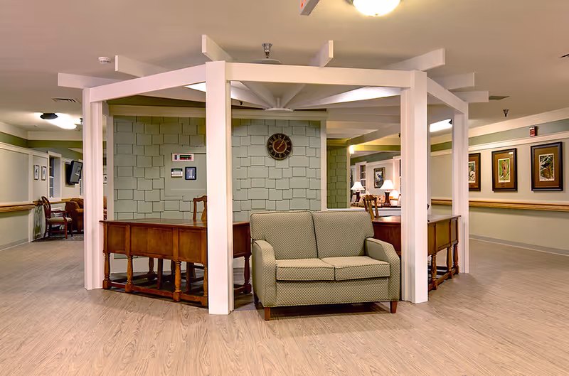 A common area in a senior living facility featuring a green patterned loveseat in front of a wooden desk structure with white beams overhead. The walls are light green with framed artwork and a clock, and the floor is light wood. There are additional chairs and lamps visible in the background along the hallway.