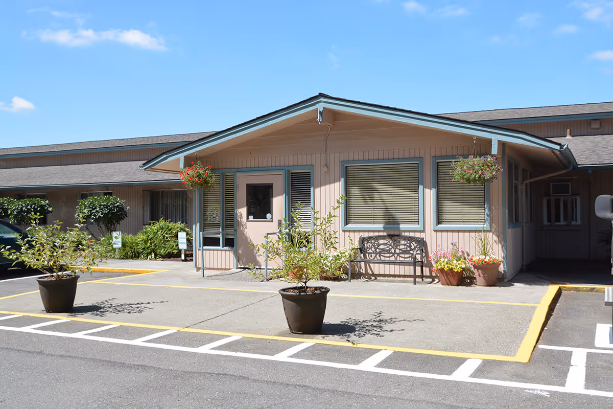 Exterior view of Benson Heights Rehabilitation Center showing a single-story building with a covered entrance. There are potted plants and hanging flower baskets near the entrance, a bench under the windows, and a parking area with marked spaces in front of the building under a clear blue sky.