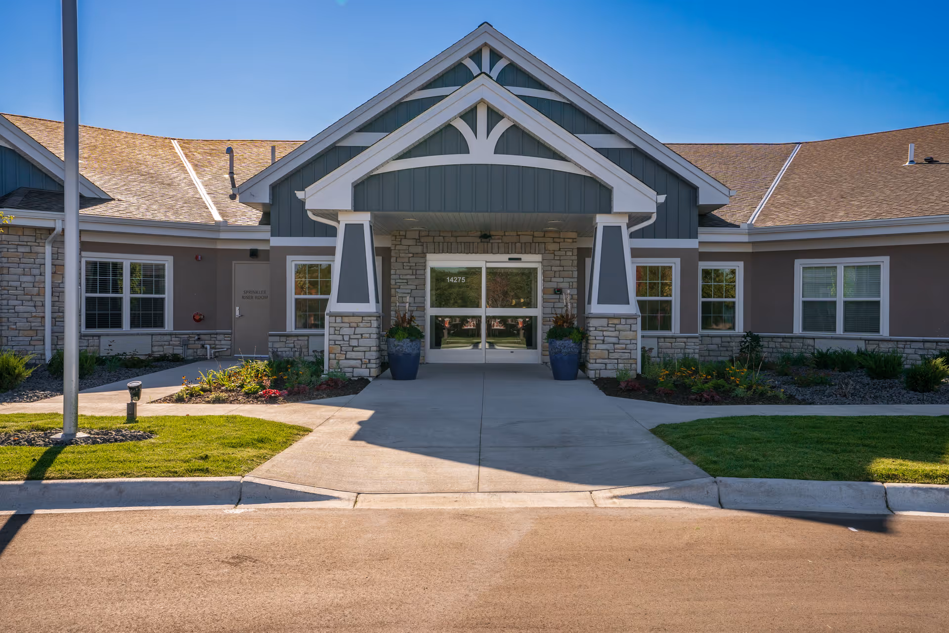 Front exterior view of NorBella Senior Living - Savage building with a covered entrance supported by stone and white pillars, large glass double doors, and landscaped greenery on either side of the walkway.