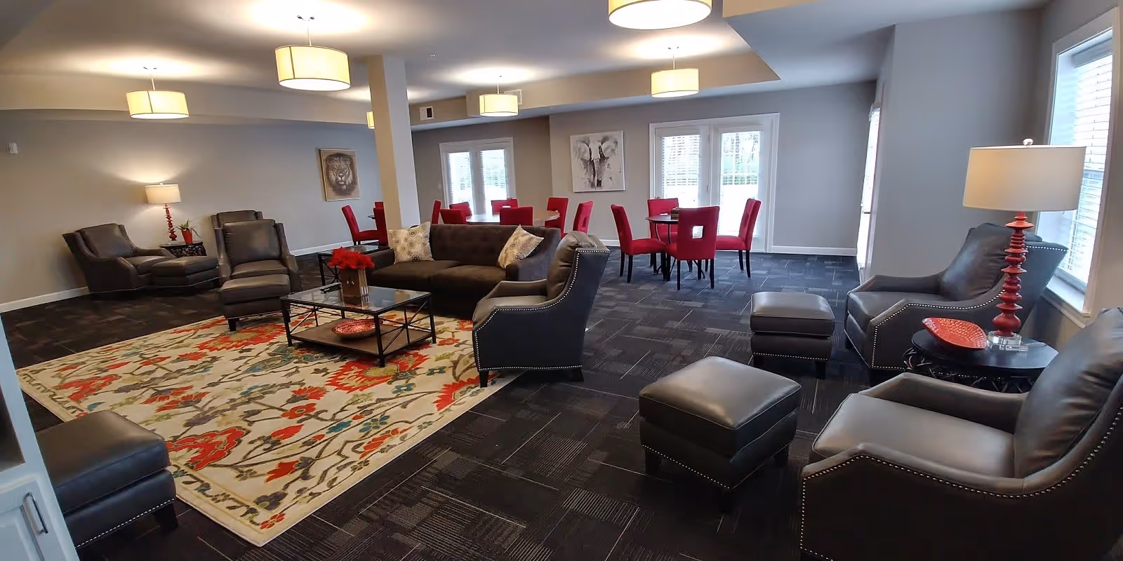 A spacious living room area with multiple dark leather armchairs and ottomans arranged around a colorful floral area rug. There is a dark sofa with decorative pillows and a glass coffee table with a red flower centerpiece. In the background, there are several red upholstered dining chairs around tables near large windows letting in natural light. The walls are light gray with framed artwork and several lamps providing warm lighting.