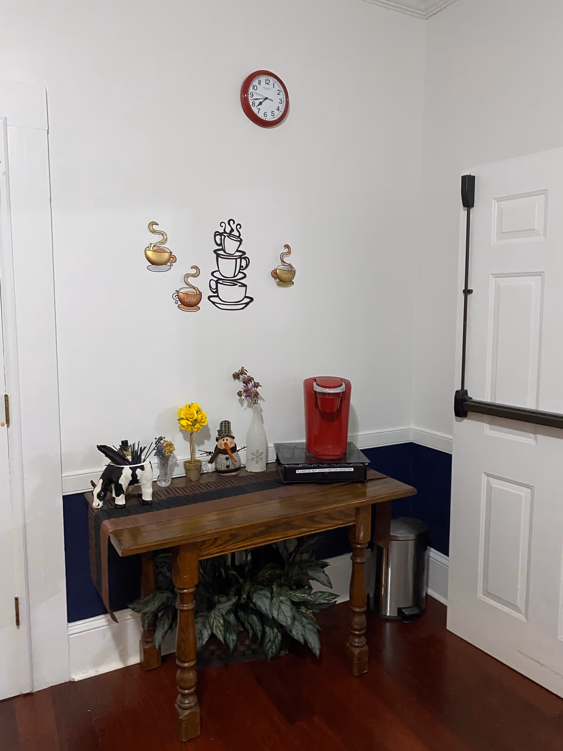 A corner of a room with a wooden table against the wall. On the table, there is a red coffee maker, a small decorative snowman, a yellow flower in a pot, a small vase with purple flowers, and a black and white cow figurine. Above the table on the wall, there are decorative images of steaming coffee cups and a round clock with a red frame showing the time as 9:35. There is a white door with a black horizontal handle on the right side and a small trash can next to the table. The lower half of the wall is painted dark blue with white trim, and the floor is wooden.