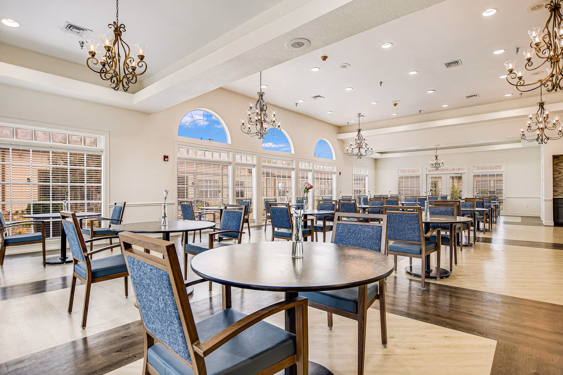 A spacious dining room with multiple round tables and blue cushioned chairs. Each table has a small vase with a single flower. The room features large windows with blinds, arched transom windows, and elegant chandeliers hanging from the ceiling. The floor has a light wood pattern with darker wood accents.