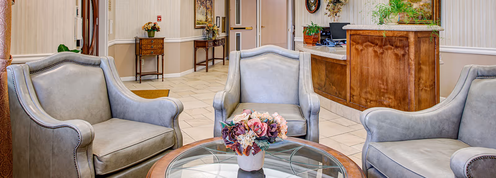 A cozy seating area in a senior living facility featuring three gray upholstered armchairs arranged around a round glass-top wooden coffee table with a floral centerpiece. In the background, there is a wooden reception desk with plants and a computer, and a hallway with decorative tables and framed artwork on the walls.