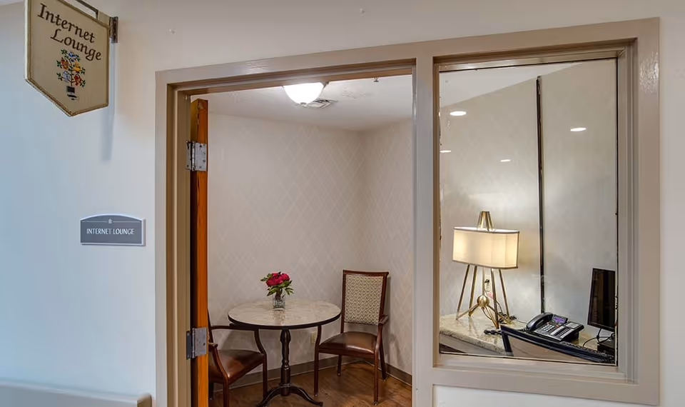 Small internet lounge room with a round table, two chairs, a desk with a lamp, a telephone, and a computer monitor. The room has light-colored wallpaper with a subtle pattern and wood flooring. A sign outside the room reads 'Internet Lounge'.