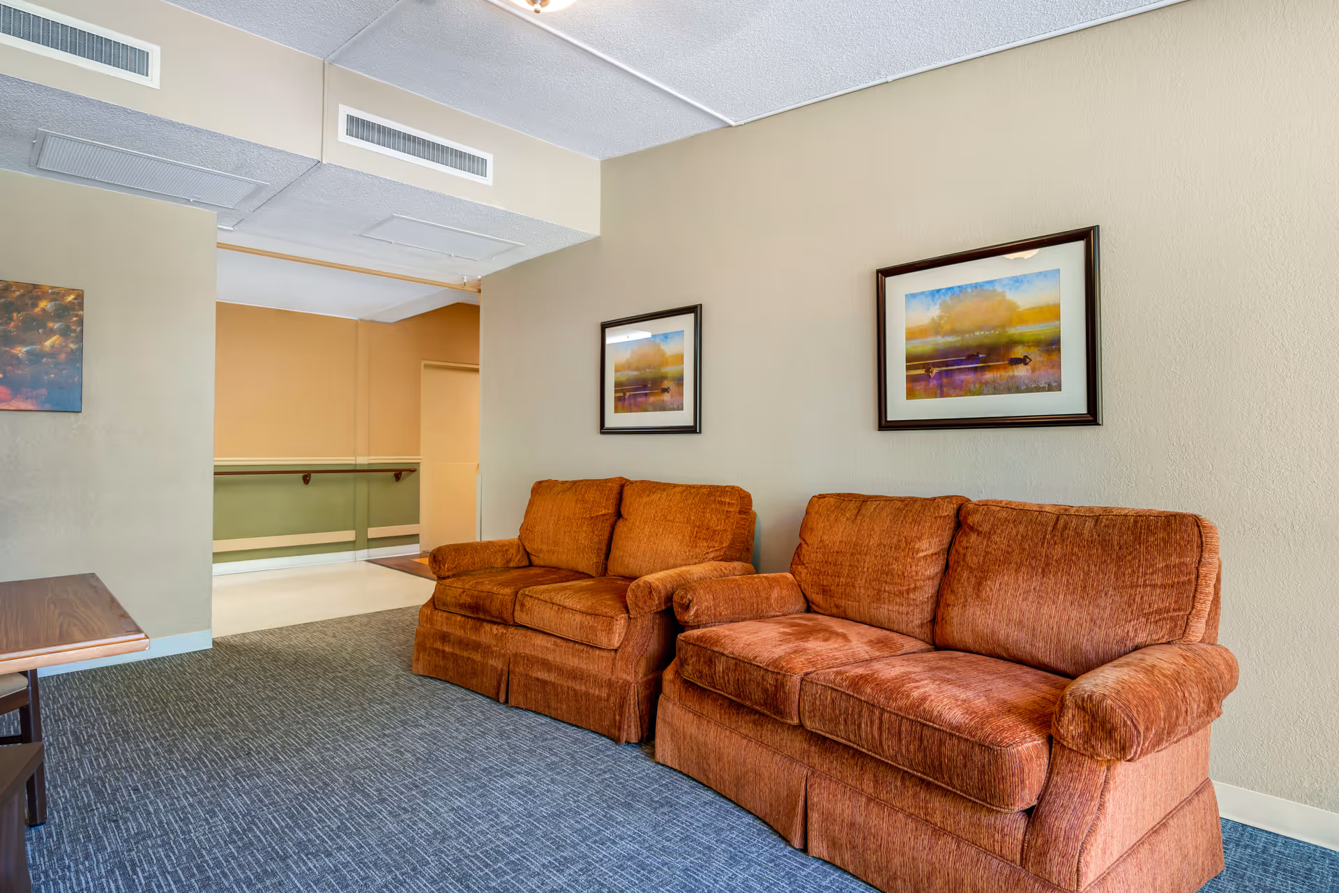 A cozy seating area in a senior living facility featuring two rust-colored upholstered sofas against a beige wall. Above the sofas are two framed landscape paintings. The room has a blue carpet and an open doorway leading to a hallway with handrails.