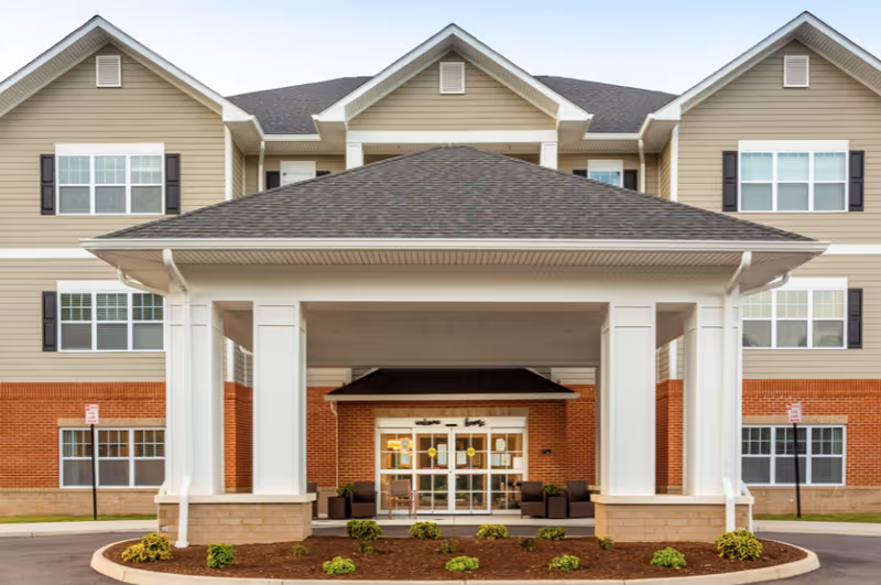 Front entrance of a multi-story senior living facility with a covered porte-cochère, brick lower facade, and windows above.