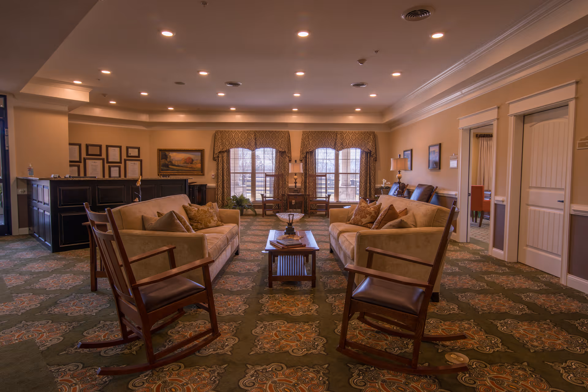 A cozy living room area in a senior living facility with two beige sofas facing each other, two wooden rocking chairs, a coffee table in the center, and large windows with patterned valances letting in natural light. The room has a patterned carpet, warm lighting from recessed ceiling lights, and framed artwork on the walls. There is a reception desk on the left side and doorways leading to other rooms on the right.