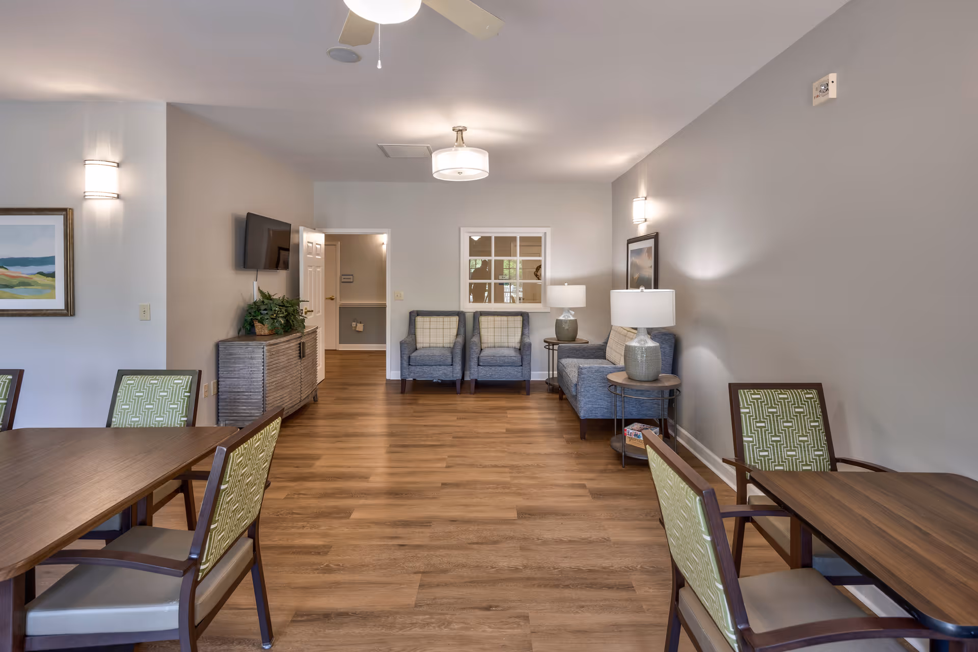A well-lit common area in an assisted living facility featuring wooden flooring, a flat-screen TV mounted on the wall, two blue armchairs, a blue sofa with two table lamps on either side, and several wooden tables with green patterned chairs. The walls are painted light gray with framed artwork and modern light fixtures.