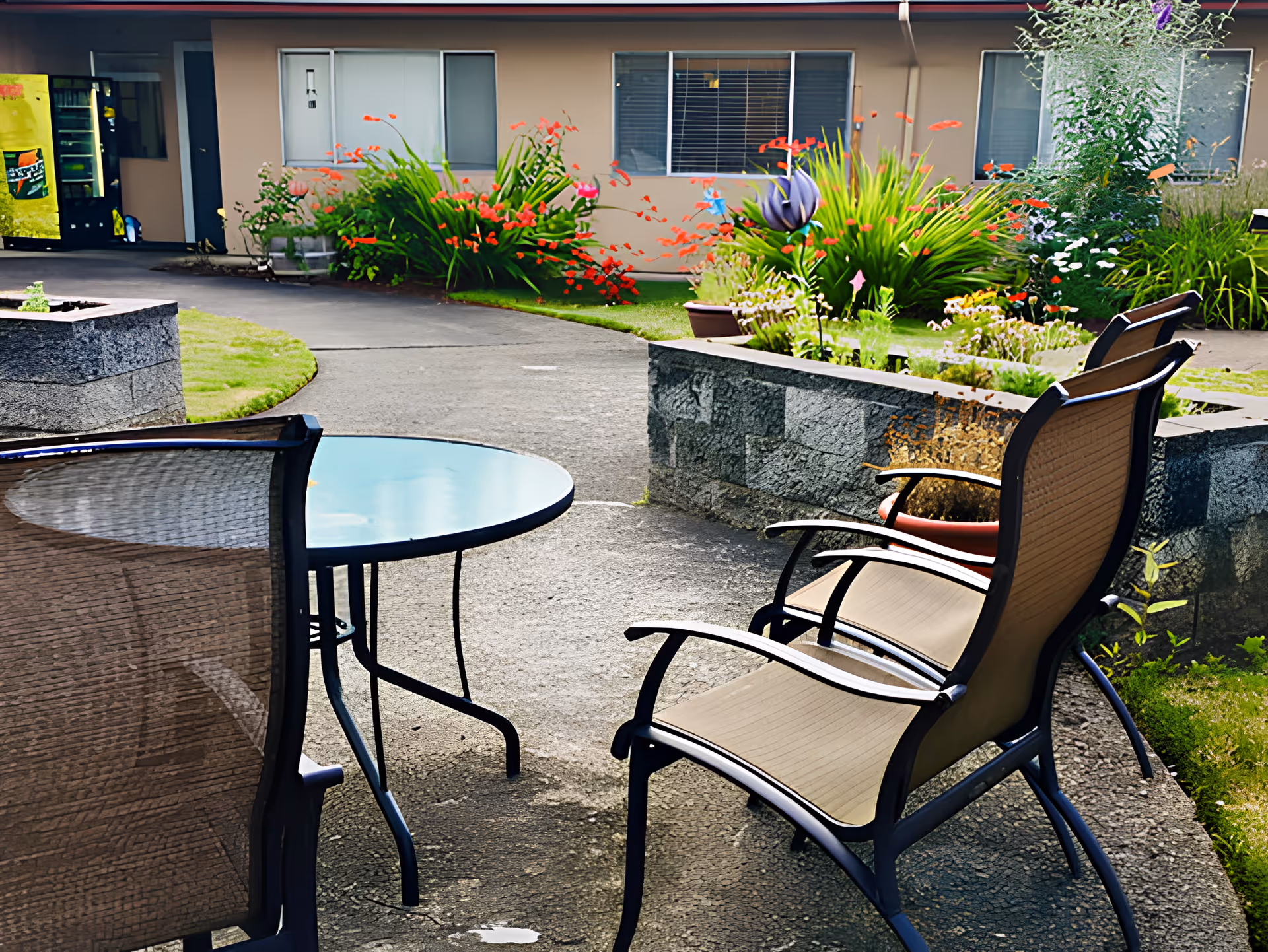 Outdoor patio area with two beige mesh chairs and a round glass-top table on a concrete surface. In the background, there are flower beds with green plants and red flowers, a vending machine, and a building with windows and a door.