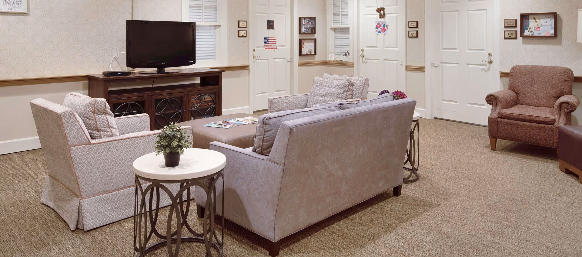 A tidy community living room with a sofa and armchairs arranged around a coffee table, a TV on a console, and several closed doors in the background.