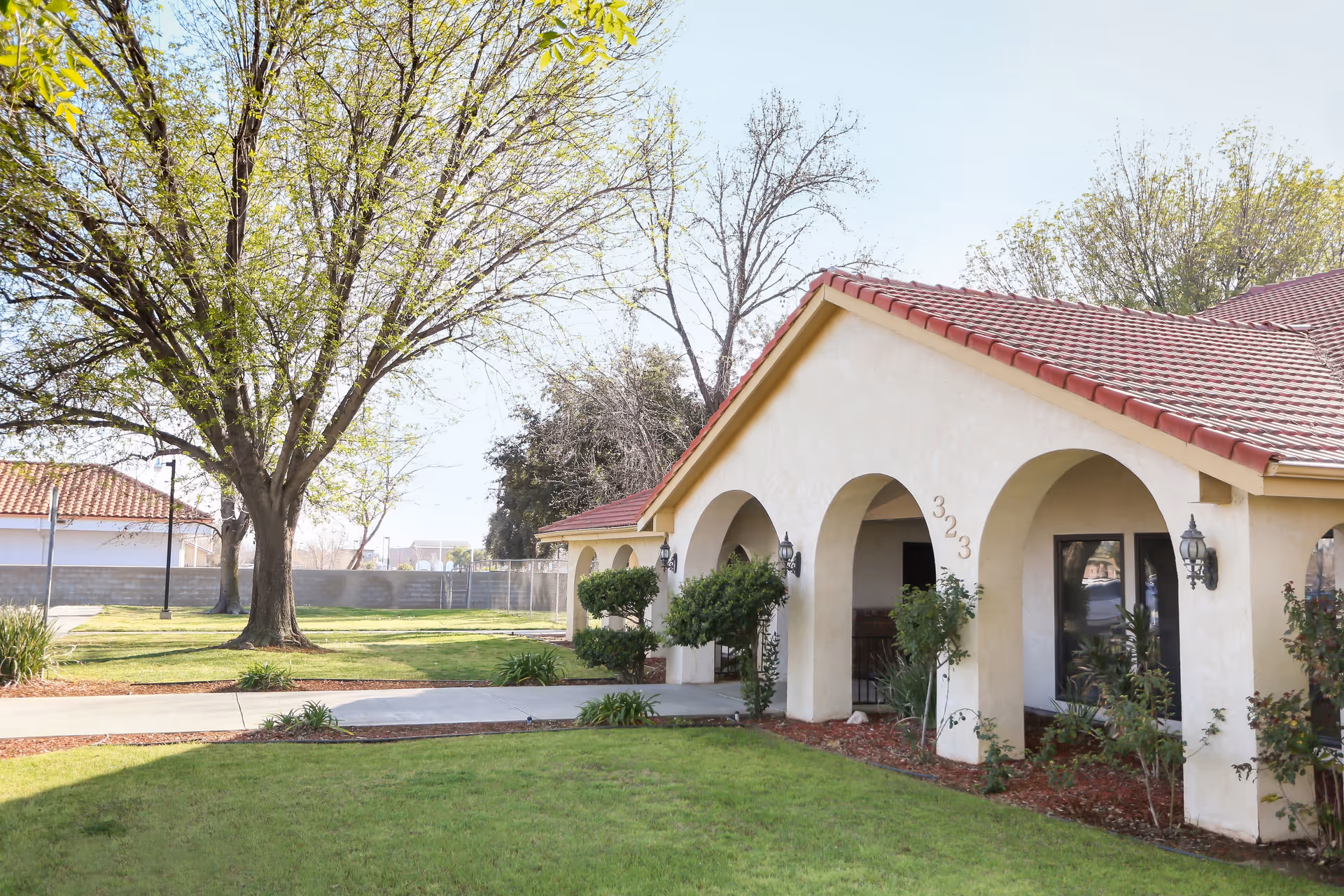 Exterior view of a single-story building with a red tile roof and beige stucco walls featuring arched doorways. The building is surrounded by a well-maintained lawn, bushes, and trees with some leaves. A concrete walkway leads to the entrance, and the number 323 is visible on the wall near the arches.