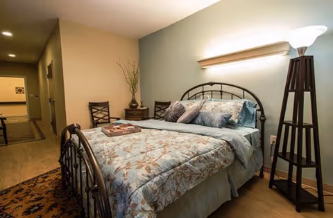 A cozy bedroom with a metal-framed bed dressed in a floral patterned comforter and multiple pillows. There is a wooden nightstand with a decorative vase and two chairs against the wall. A tall floor lamp with a white shade stands beside the bed, and a hallway is visible in the background.