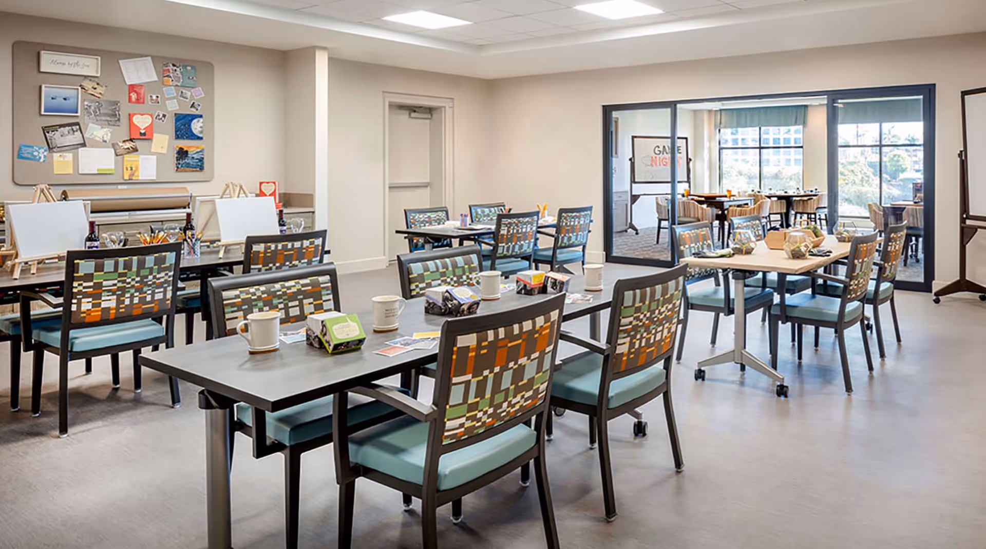 A bright, spacious activity room with multiple tables and chairs arranged for group activities. The tables have art supplies, mugs, and small boxes on them. There are easels with blank canvases and a bulletin board with various notes and pictures on the wall. Large windows and glass doors reveal another room with more tables and chairs, and a whiteboard with 'Game Night' written on it.