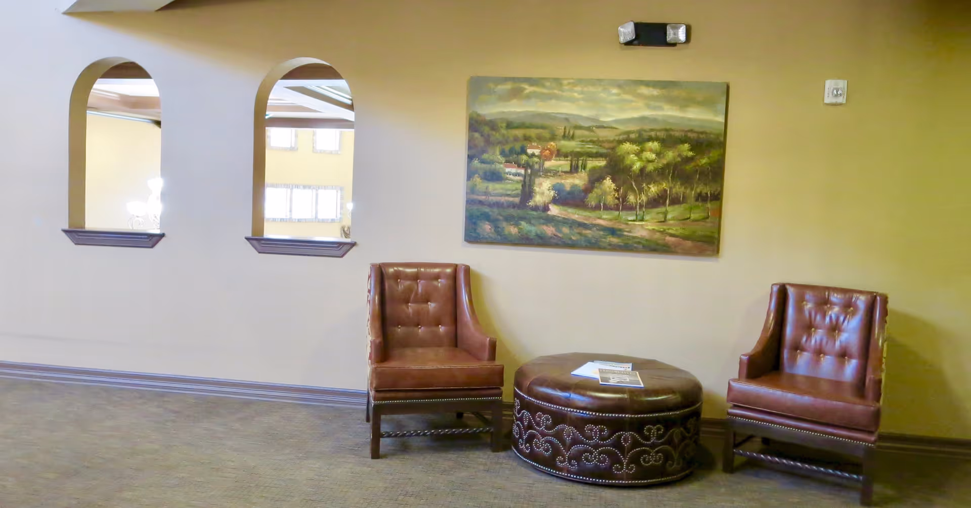 Two leather armchairs flank a round upholstered ottoman beneath a landscape painting in a quiet seating area with arched interior windows.