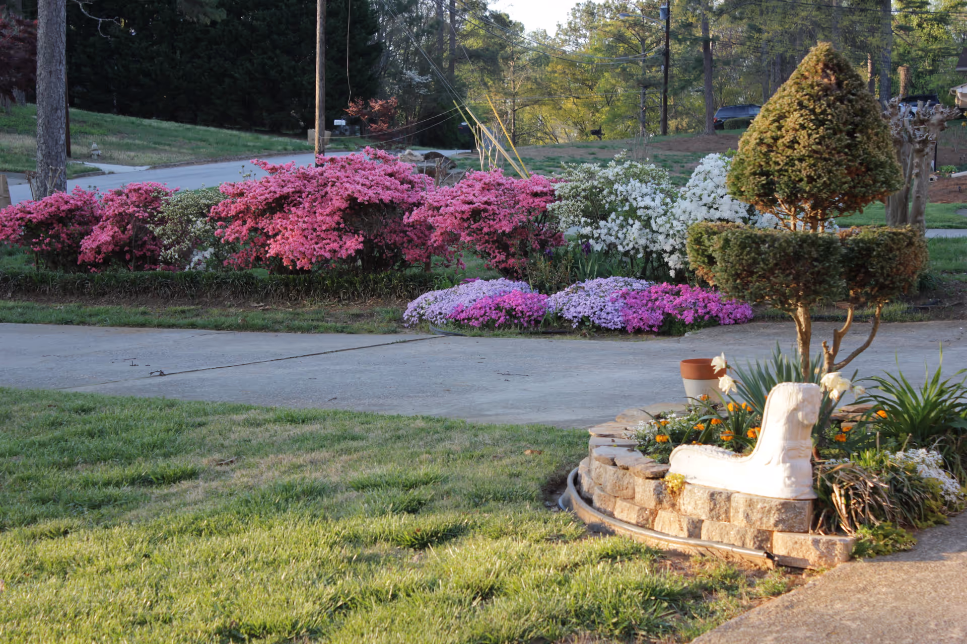 A landscaped outdoor area featuring a variety of colorful flowering bushes including pink, white, and purple flowers. There is a neatly trimmed topiary tree shaped in a tiered design planted in a circular stone planter with additional flowers and greenery. A concrete driveway or pathway runs through the scene with grass on either side, and trees and a road are visible in the background.
