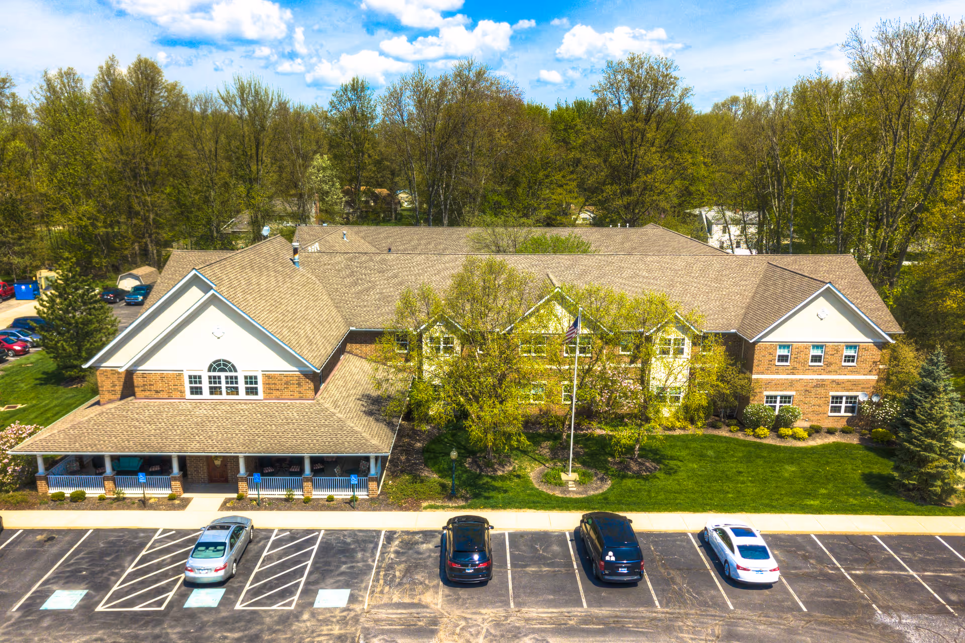 Aerial view of a two-story brick building with a large covered porch, surrounded by green trees and a parking lot with several cars parked. The building is set against a backdrop of dense trees under a partly cloudy sky.