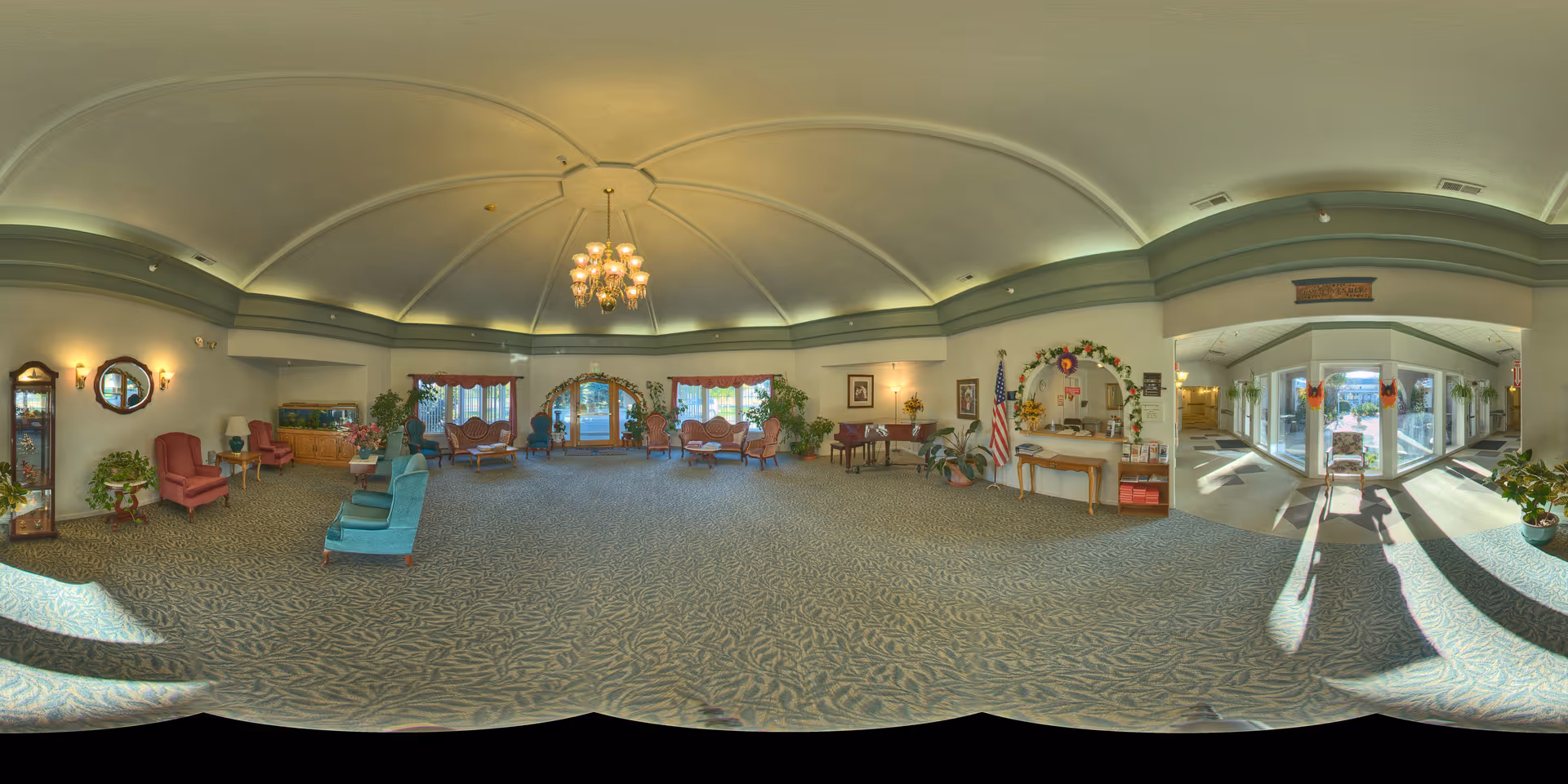A spacious and well-lit assisted living facility common area with a high, curved ceiling and a central chandelier. The room is furnished with various seating options including armchairs and sofas arranged in small groups. There are plants, framed pictures on the walls, and an American flag near a decorated archway. Large windows and glass doors allow natural light to fill the space, and a piano is visible against one wall.