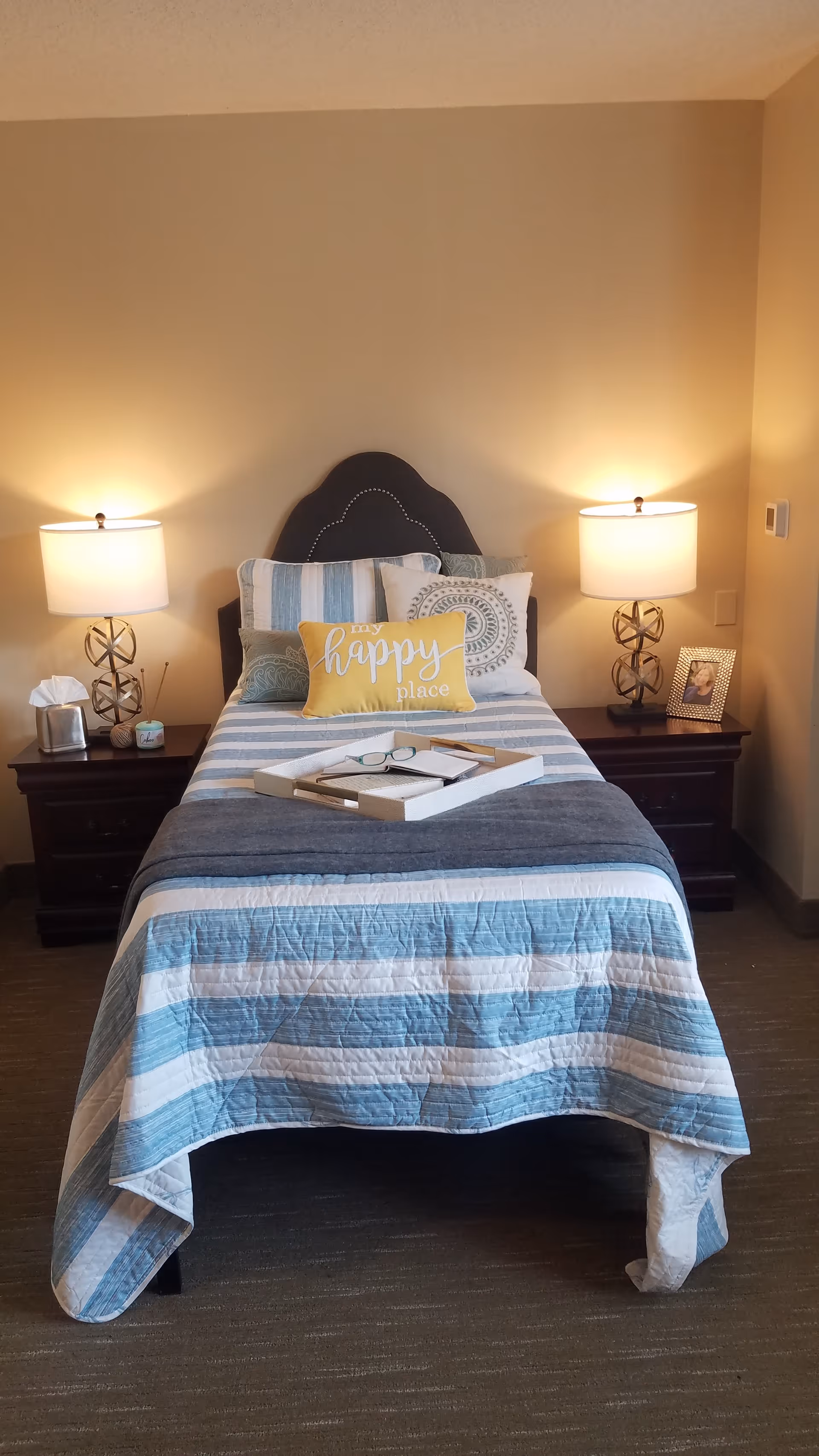 A neatly made single bed with a blue and white striped quilt and multiple pillows, including a yellow pillow with the words 'my happy place'. The bed has a dark headboard and is flanked by two dark wooden nightstands, each with a modern lamp. One nightstand has a tissue box and a small candle, while the other has a framed photo. A tray with a book and glasses rests on the bed.