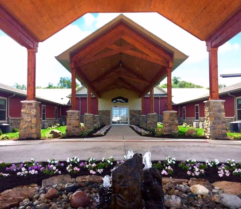 Entrance of Copper Creek Cottages Memory Care of Mattoon featuring a covered walkway with wooden beams and stone pillars. In the foreground, there is a small rock water fountain surrounded by flower beds with purple and white flowers. The building has red siding and stone accents.