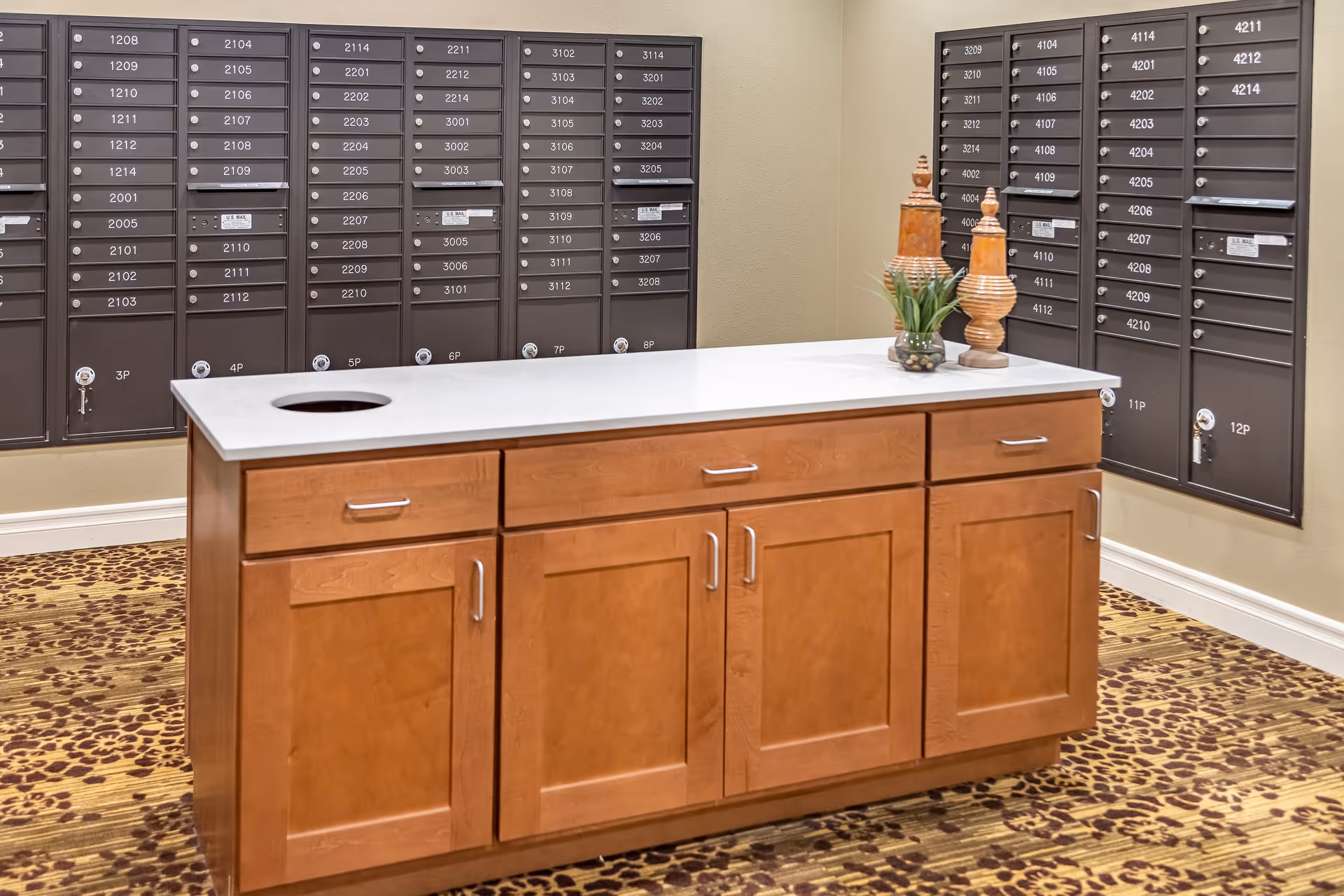 Interior mailroom with multiple rows of dark brown mailboxes mounted on beige walls. In the center, there is a wooden cabinet with a white countertop, decorated with two wooden ornamental pieces and a small potted plant. The floor is covered with a patterned carpet in shades of brown and beige.